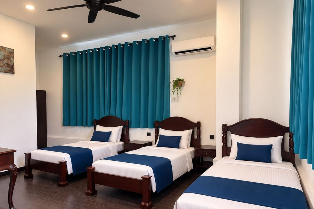 Une chambre d’hôtel avec trois lits en bois, un ventilateur de plafond et des rideaux de couleur turquoise.