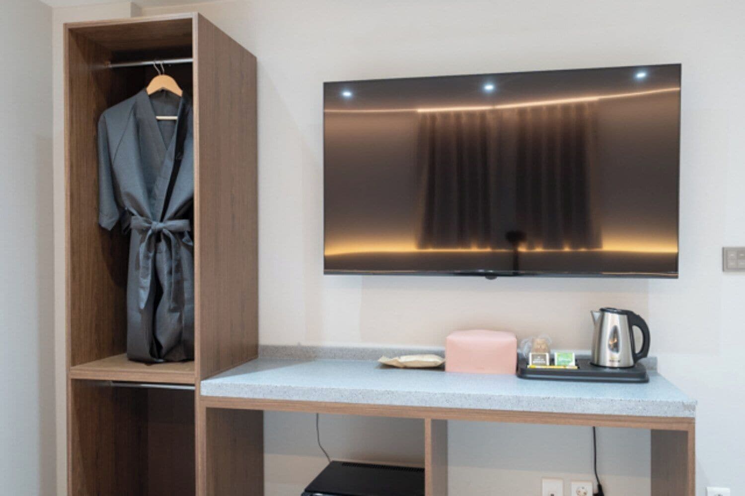 A room with a wooden wardrobe, a flat-screen TV mounted on the wall, a small table with a kettle and a pink candle.