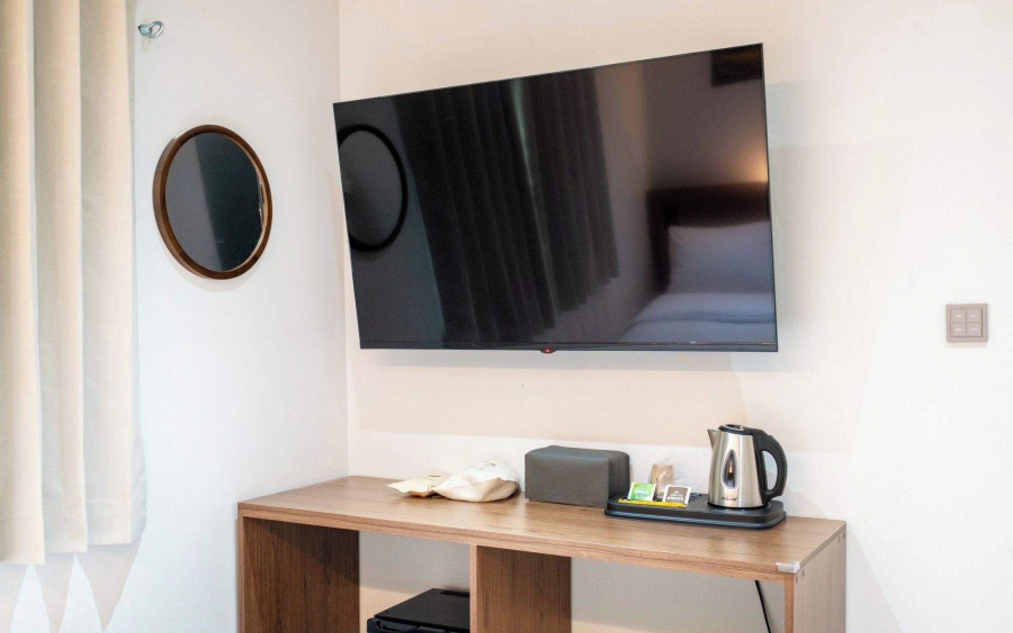 A modern hotel room with a flat-screen TV mounted on the wall, a wooden desk with a kettle and a tray of items, and a round mirror on the wall.