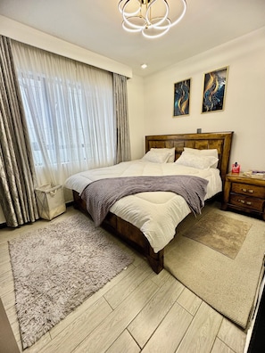 A bedroom with a wooden bed, a nightstand, a lamp, a window with curtains, and two wall art pieces.