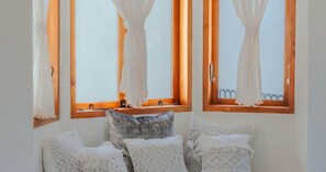 Ein Zimmer mit drei Fenstern, jeweils mit Holzrahmen und weißen Vorhängen. Auf einer hölzernen Bank liegen mehrere weiße Kissen und ein blaues Sitzkissen.