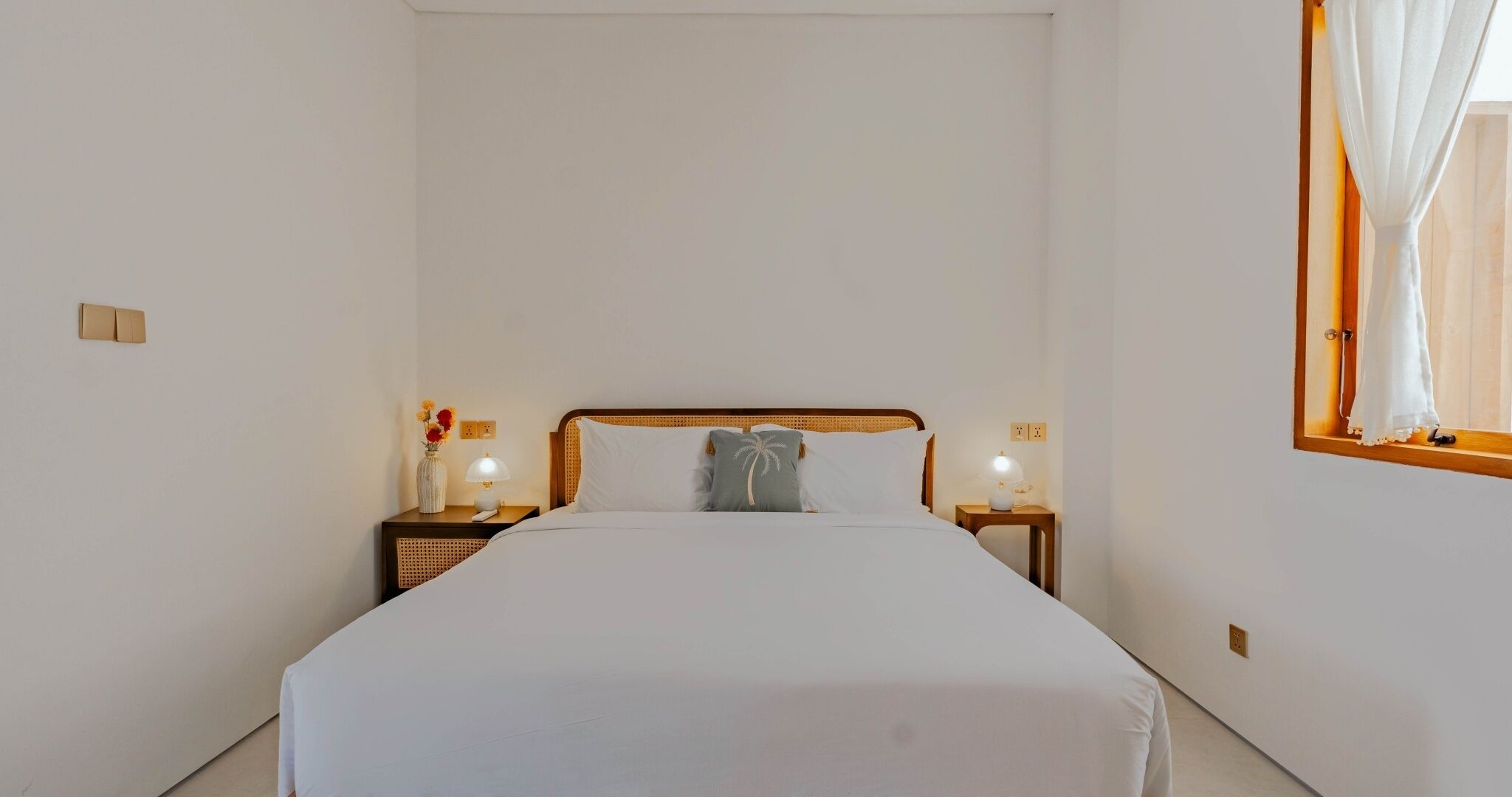 A hotel room with a single bed, two bedside tables with lamps, a window with curtains, and a wall socket.