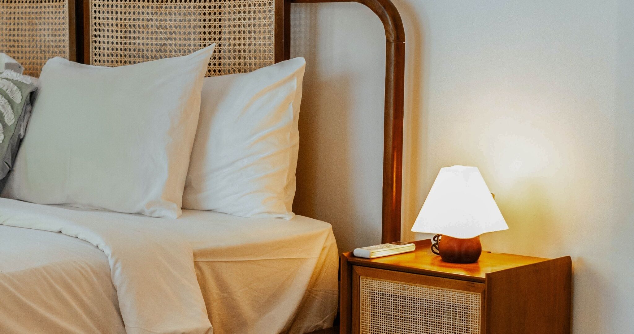 Una cama bien hecha con sábanas blancas, cabecera de madera y mesita de noche con una lámpara y un control remoto.