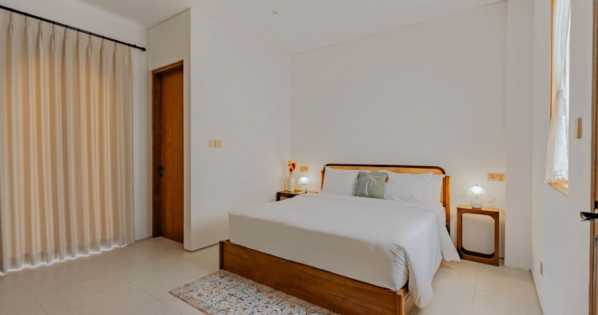 A bedroom with a wooden bed, two bedside tables with lamps, a door, and a window with curtains.