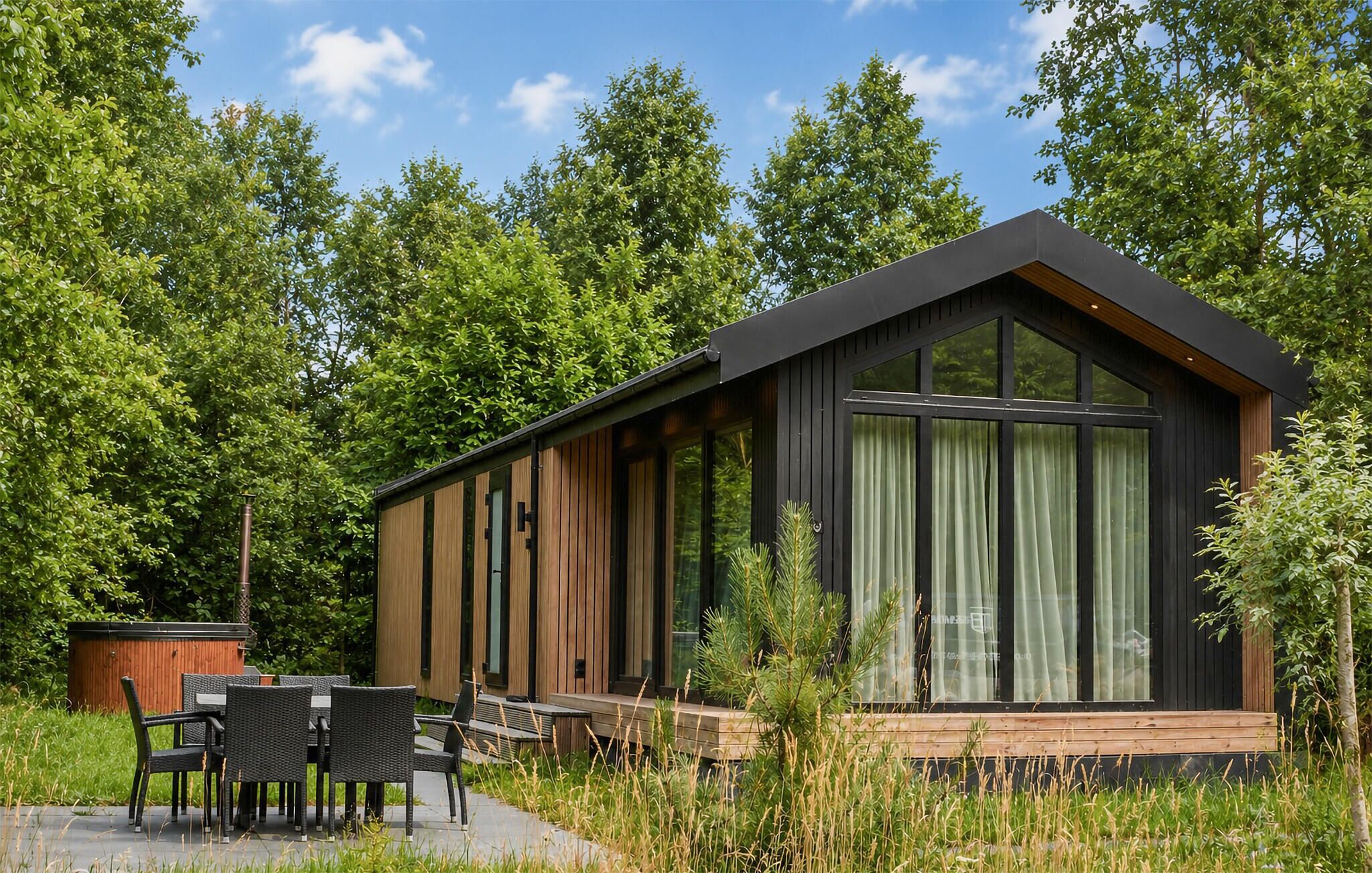 Una cabaña moderna con un ventanal grande, una terraza de madera y un comedor con mobiliario exterior.