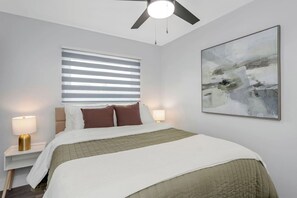 Een hotelkamer met een bed, een plafondventilator, een raam met zonwering, een bedlamp, een wanddecoratie met abstracte kunst en een nachtkastje.