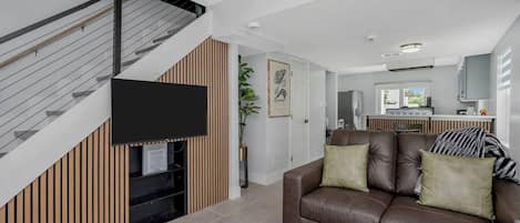 A modern living room with a brown leather sofa, a flat-screen TV mounted on a wall, and a staircase with a metal railing.