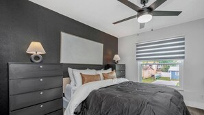 Ein Schlafzimmer mit einem dunkelbraunen Bett, einem Deckenventilator, einem Fenster mit Jalousien und einem Nachttisch mit Lampe.