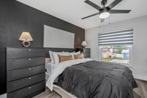 Un dormitorio con cama oscura, ventilador de techo, ventana con persianas y mesita de noche con lámpara.