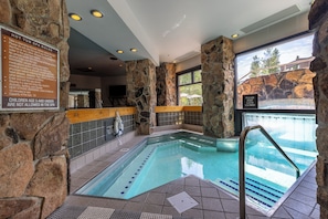 An indoor swimming pool with stone walls and a wooden deck, surrounded by windows. - Ski-in/ski-out! Big Sky Resort! Community Pool/Hot Tub! (Big Sky)