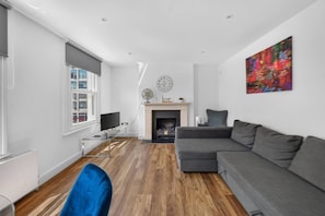 A modern living room with a grey sofa, a fireplace, a clock, and a colorful abstract painting. - Stylish London Flat Spr4 (London)