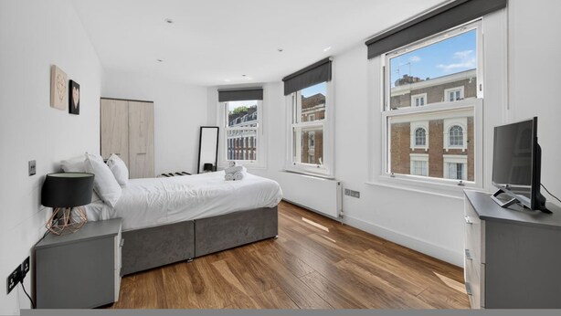 Quarto moderno com cama, mesas de cabeceira, um espelho e janelas amplas.