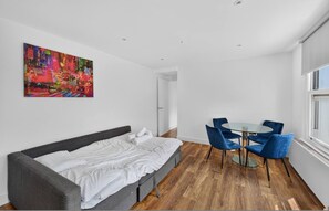 A modern bedroom with a bed, a painting, a dining area with chairs, and a window. - Stylish London Flat Spr4 (London)