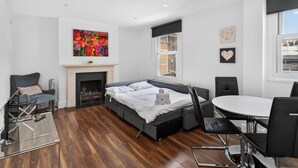 A modern bedroom with a bed, a fireplace, a dining table, and a view of the city. - Cool London Flat Spr3 (London)