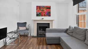A modern living room with a grey sectional sofa, a glass coffee table, a fireplace, and a colorful abstract painting above it. - Cool London Flat Spr3 (London)