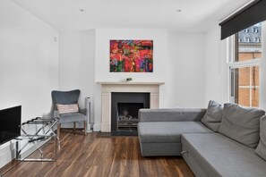 Ein modernes Wohnzimmer mit einem grauen Ecksofa, einem Glastisch, einem Kamin und einem farbenfrohen, abstrakten Gemälde darüber.