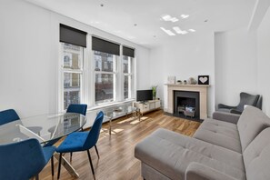 Ein modernes Wohnzimmer mit einem Glastisch, blauen Stühlen, einem grauen Sofa, einem Kamin und großen Fenstern.