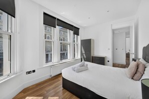 A modern bedroom with a large bed, a dresser, a mirror, and a window with a view of buildings. - Trendy London Apartment Spr2 (London)