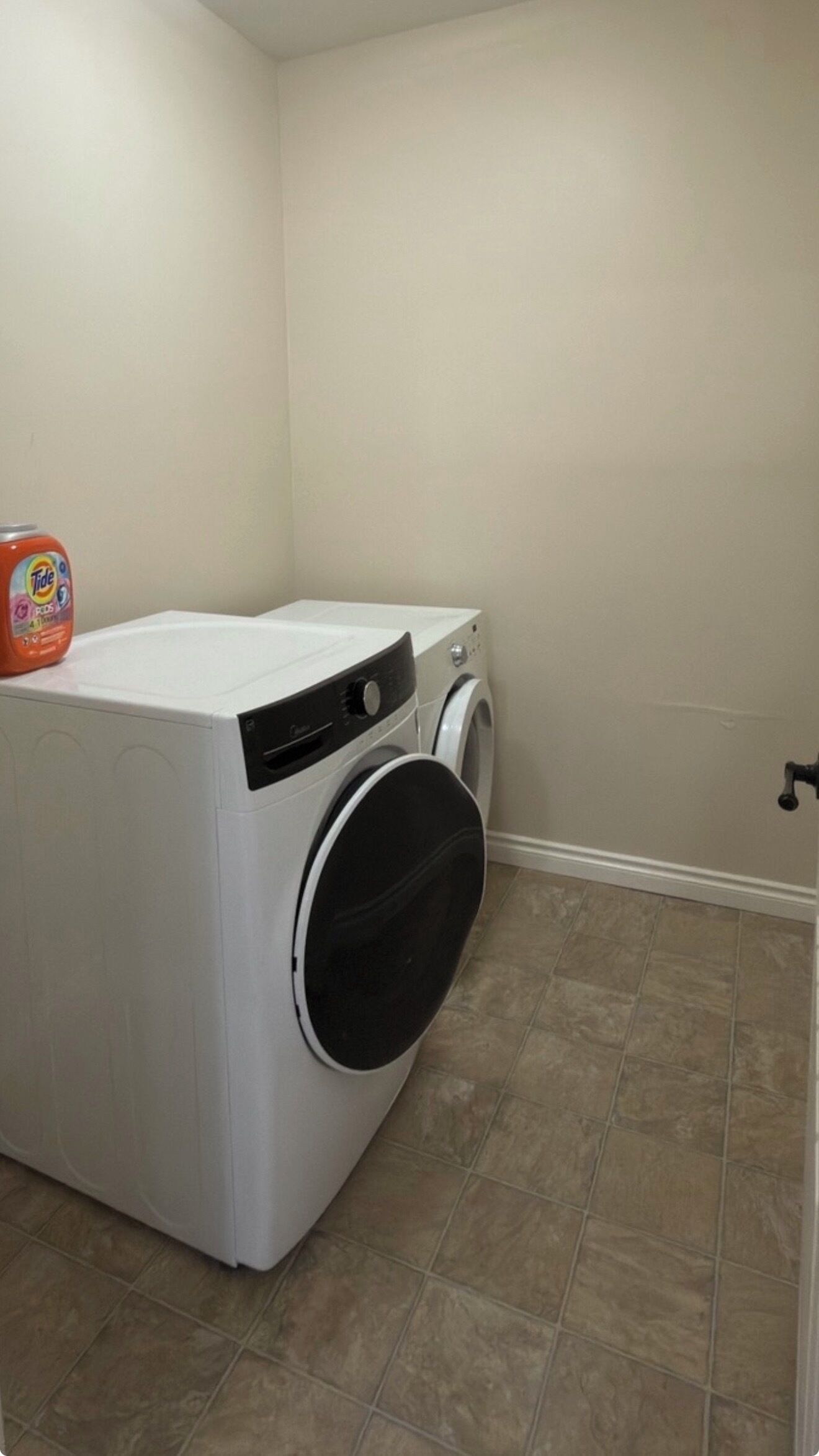 A compact, white front-loading washer and dryer unit in a corner of a room with beige walls and tiled flooring.
