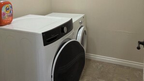 Un appareil compact, blanc, combinant une machine à laver et un sèche-linge à chargement frontal, placé dans un coin d’une pièce aux murs de couleur beige et au sol carrelé.