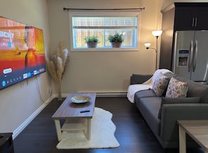 A living room with a flat-screen TV, a sofa, a coffee table, a window with plants, and a refrigerator.