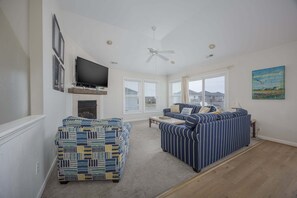 A living room with a fireplace, a television, a ceiling fan, and a large window with blinds. - 5433 - Sonnie Beaches Ocean-view 4BR, 4.5BA home with private pool, hot tub, and game room. (Nags Head)