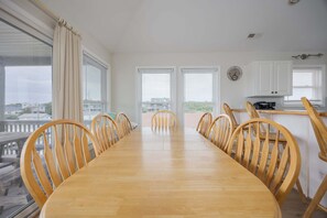 A dining room with a long wooden table and chairs, large windows with blinds, and a view of a balcony. - 5433 - Sonnie Beaches Ocean-view 4BR, 4.5BA home with private pool, hot tub, and game room. (Nags Head)