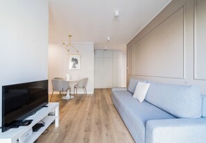 A modern living room with a flat-screen TV, a grey sofa, a dining area with a white table and chairs, and a hallway leading to a wardrobe. - Sikorskiego Serene Apartment (Poznań)