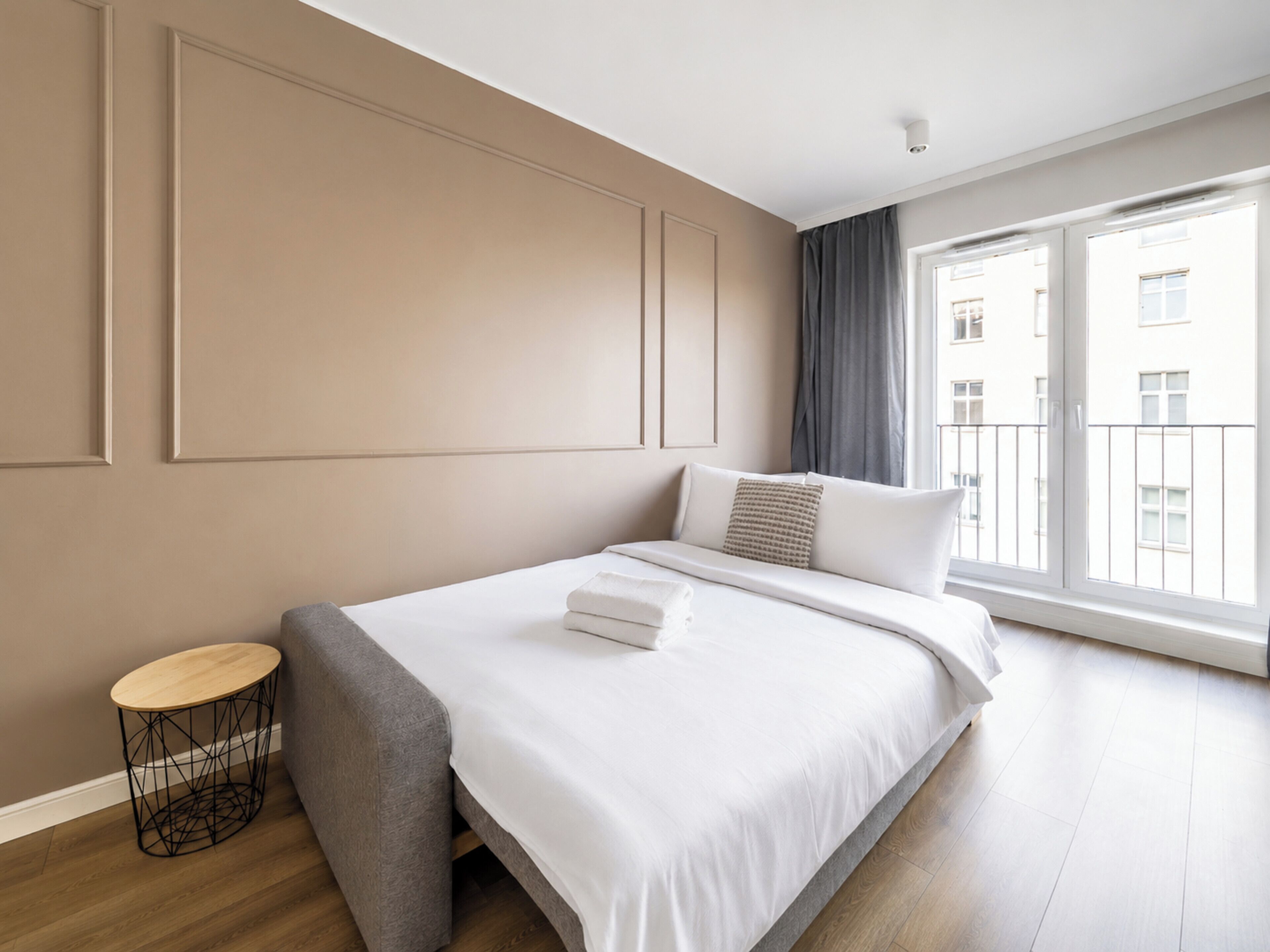 A modern bedroom with a large bed, a round side table, and a view of the city through a large window.