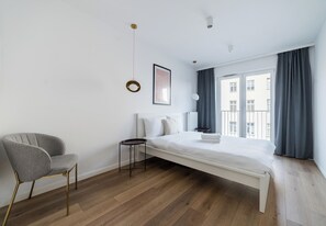 A modern bedroom with a bed, a chair, a bedside table, a framed picture, and a window with curtains. - Sikorskiego Serene Apartment (Poznań)
