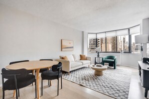 A modern living room with a dining table, sofa, and chairs, a patterned rug, and a view of buildings through large windows. - Blueground | Manhattan, rooftop, nr Empire State (New York)