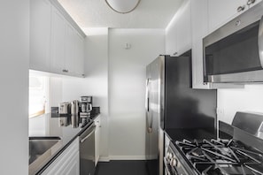 A compact kitchen with white cabinets, a stainless steel refrigerator, a gas stove, and a coffee maker. - Blueground | Manhattan, rooftop, nr Empire State (New York)
