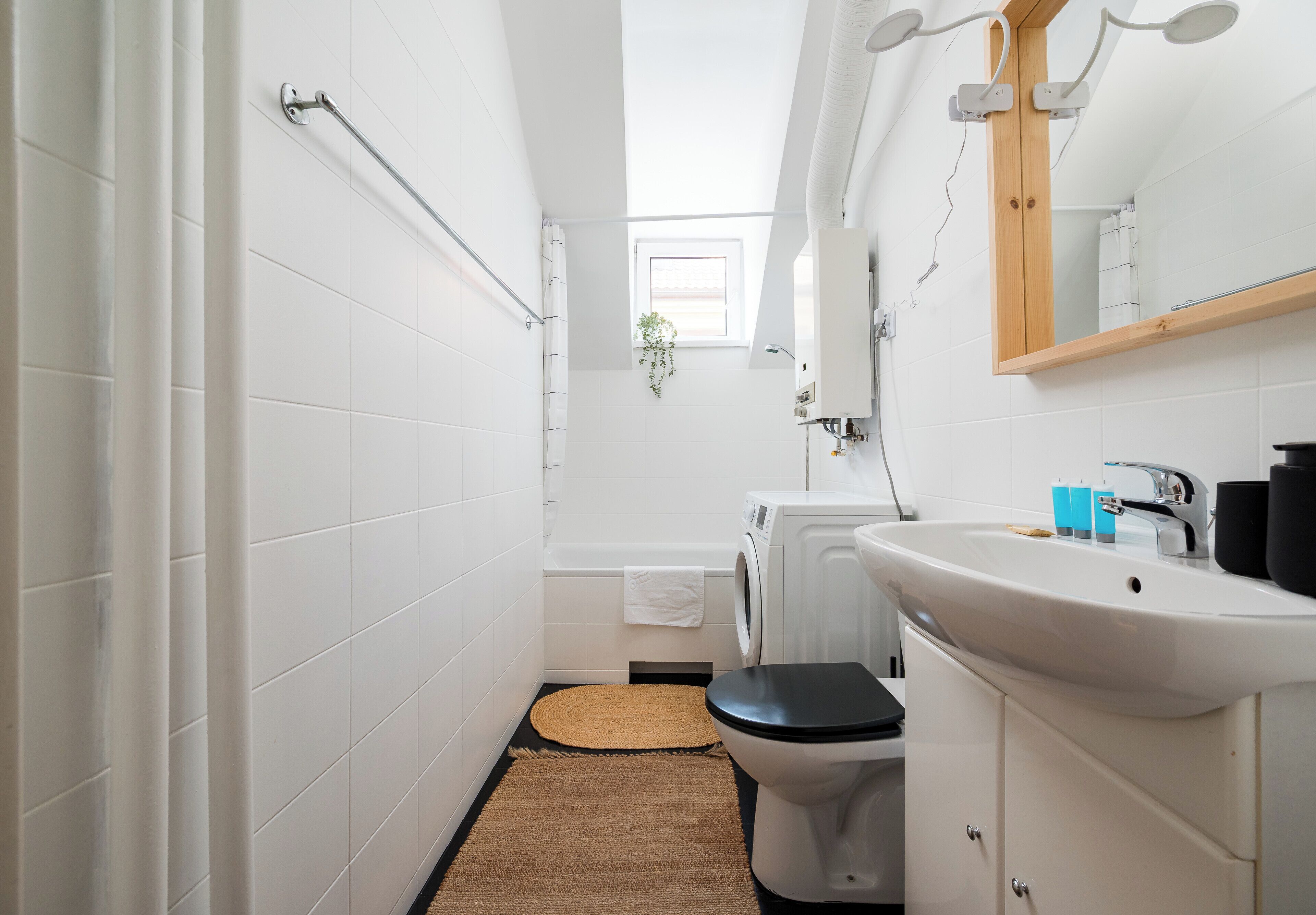Ein modernes Badezimmer mit weißen Fliesen, einer schwarzen Toilette, einer Waschmaschine und einem Waschbecken mit Spiegel darüber.