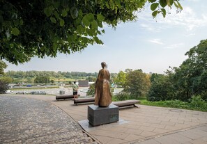 En staty på en belagd yta med bänkar, omgiven av grönska och med utsikt över en väg och byggnader i fjärran.