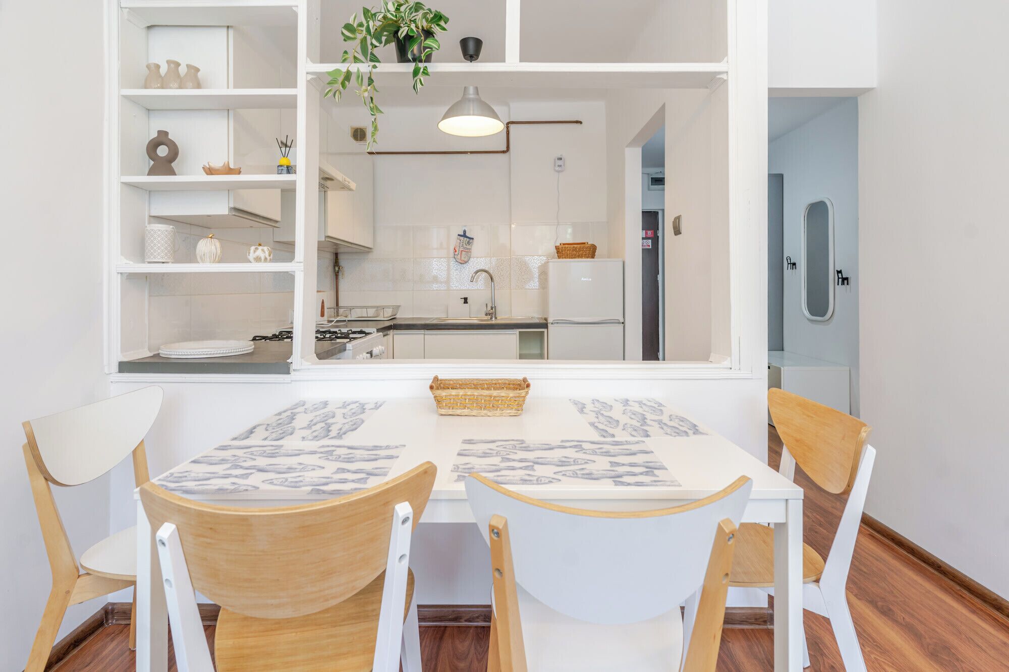 Un coin repas avec une table et des chaises blanches, une plante suspendue, et une vue sur une cuisine avec des armoires blanches et un fourneau.