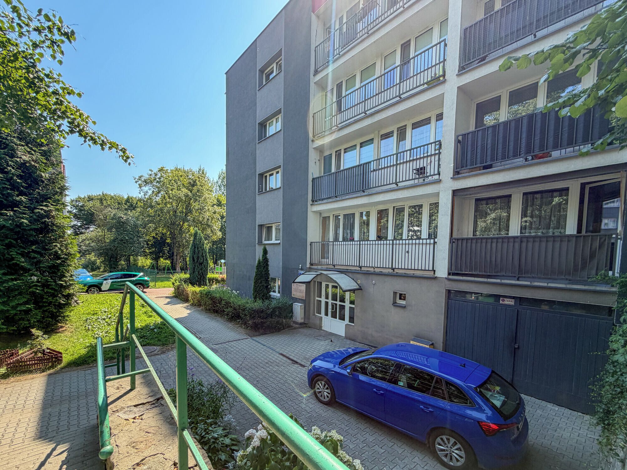 Un edificio residencial de varios pisos con balcones, una entrada con reja y un camino que lleva a un coche estacionado.