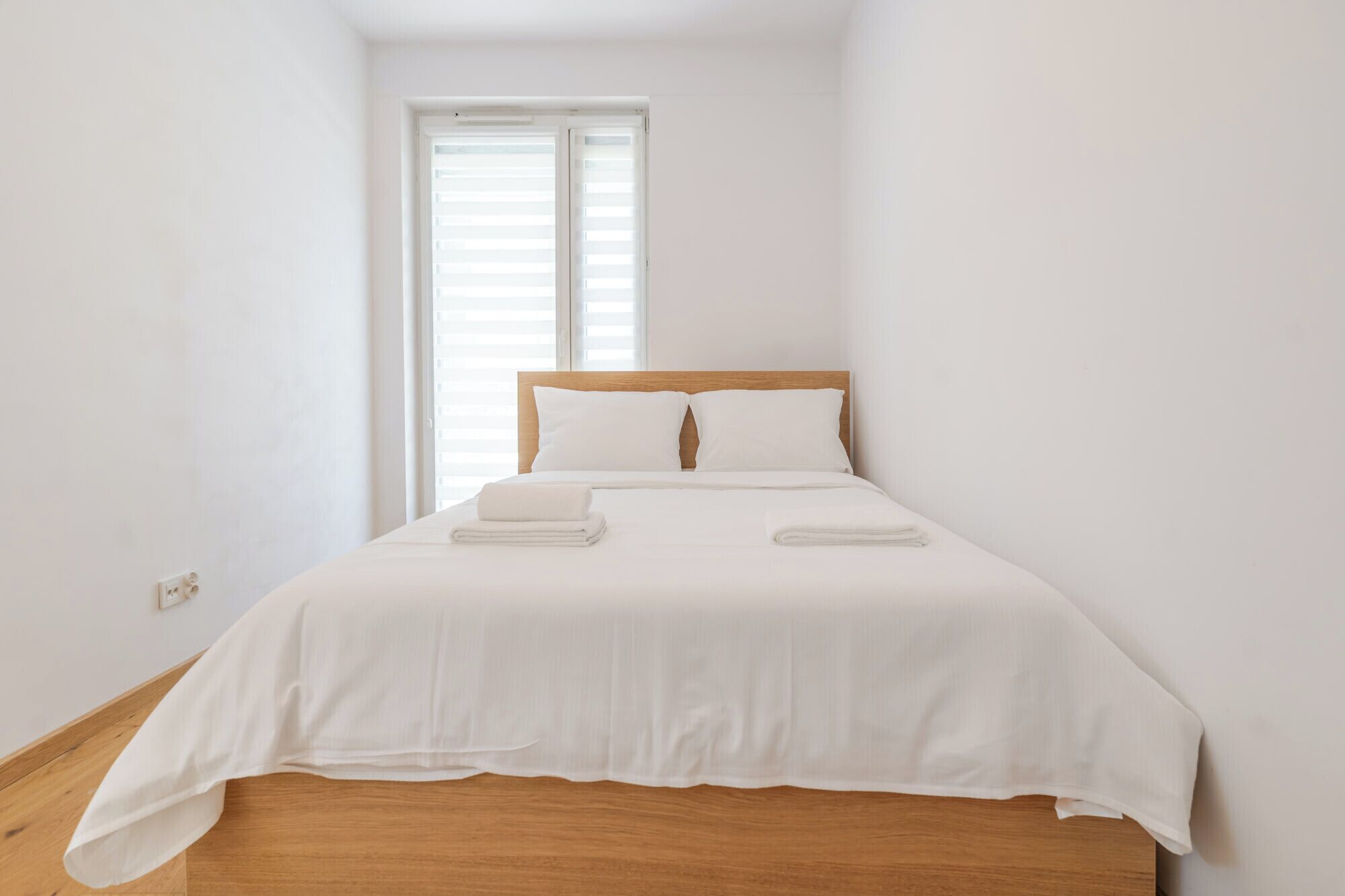 Een slaapkamer met een houten hoofdeinde, wit beddengoed en een raam met zonwering.