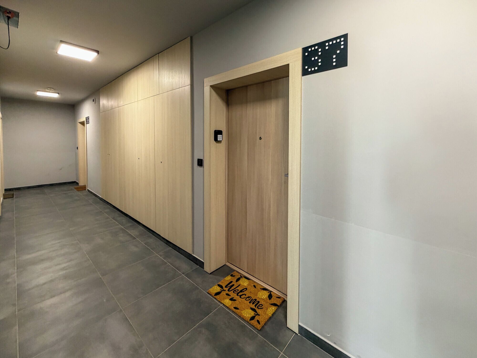 Un pasillo con una puerta de madera, un reloj digital y un felpudo.