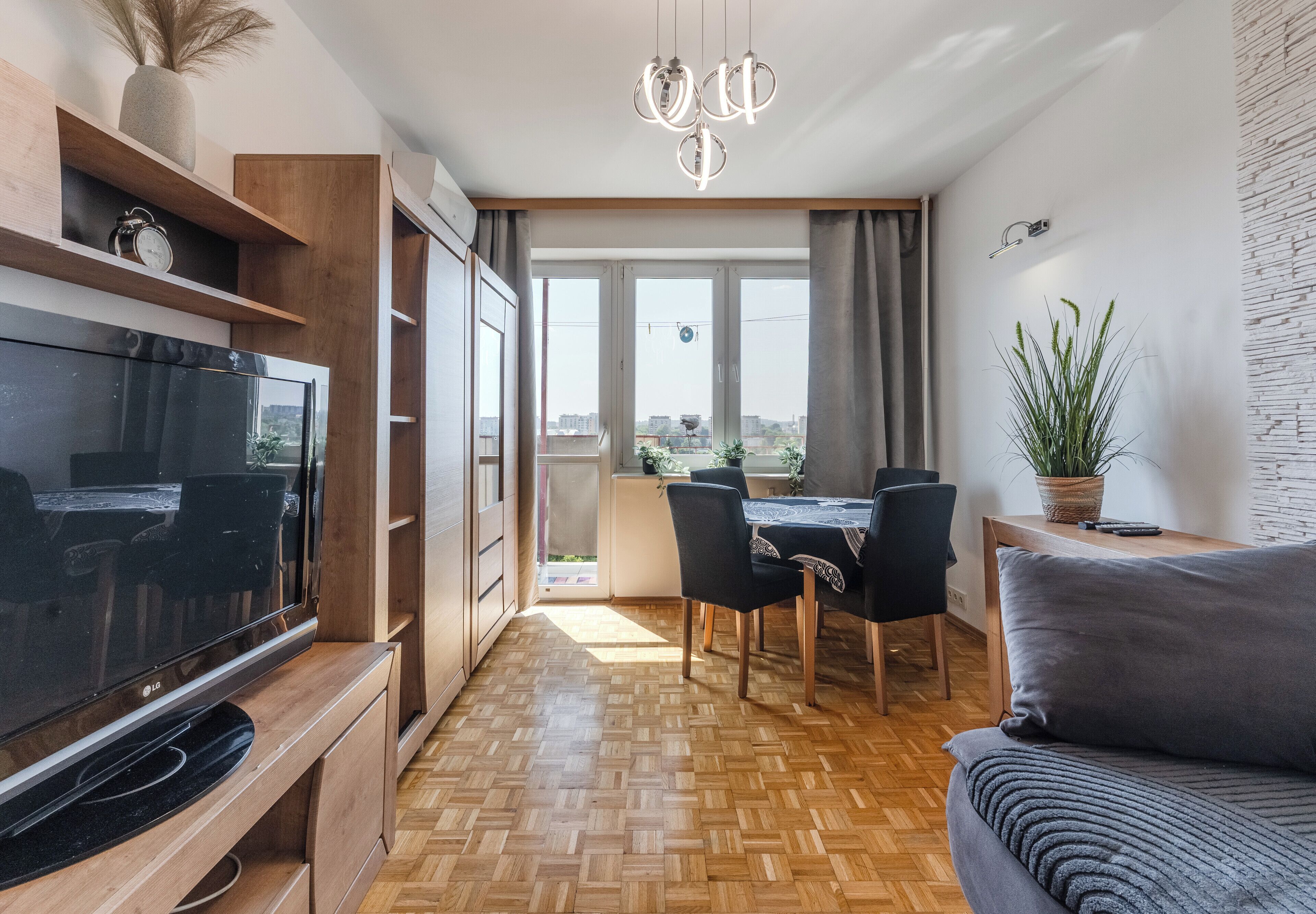 A modern living room with a flat-screen TV, wooden flooring, a dining area with chairs, and a large window offering a view of the city.