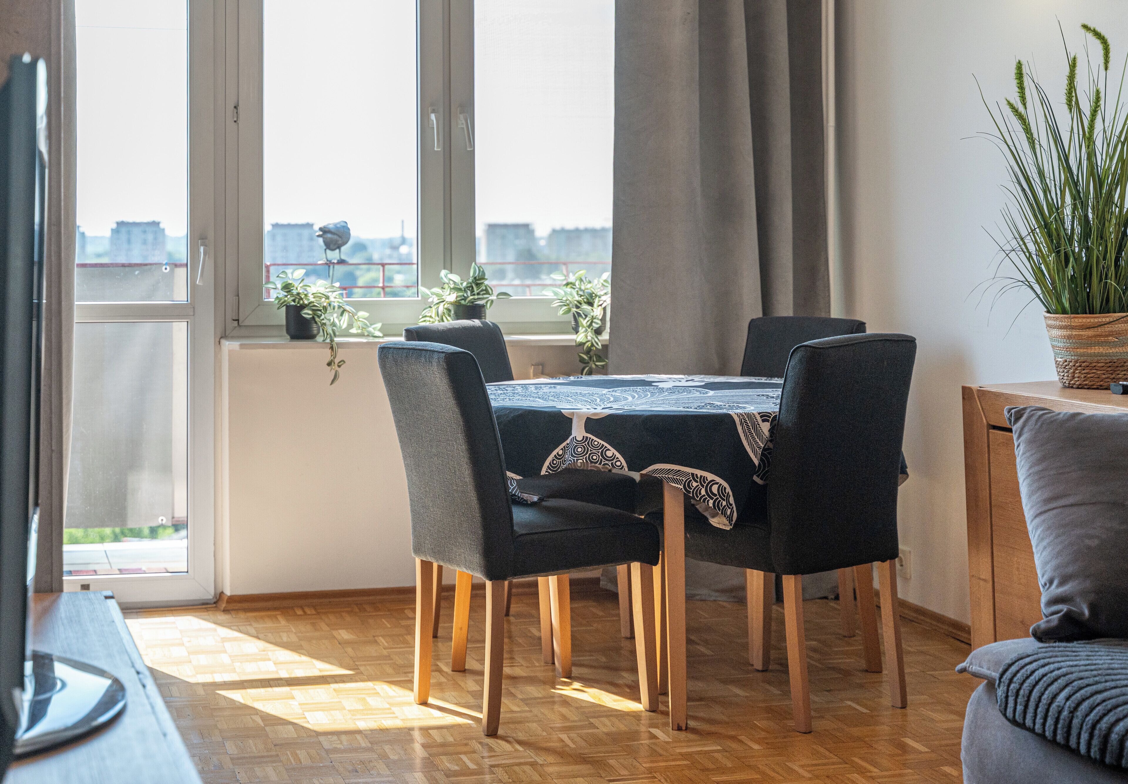 Ruokailutila, jossa on neljälle katettu pöytä, pöydän ympärillä tuoleja, ruukkukasvi ja näkymä ikkunasta.