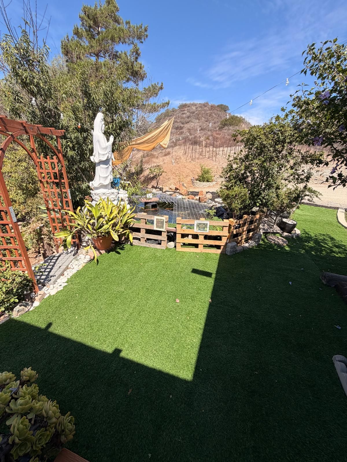 Un jardí amb gespa artificial, una tanca de fusta, una estàtua i una pèrgola.