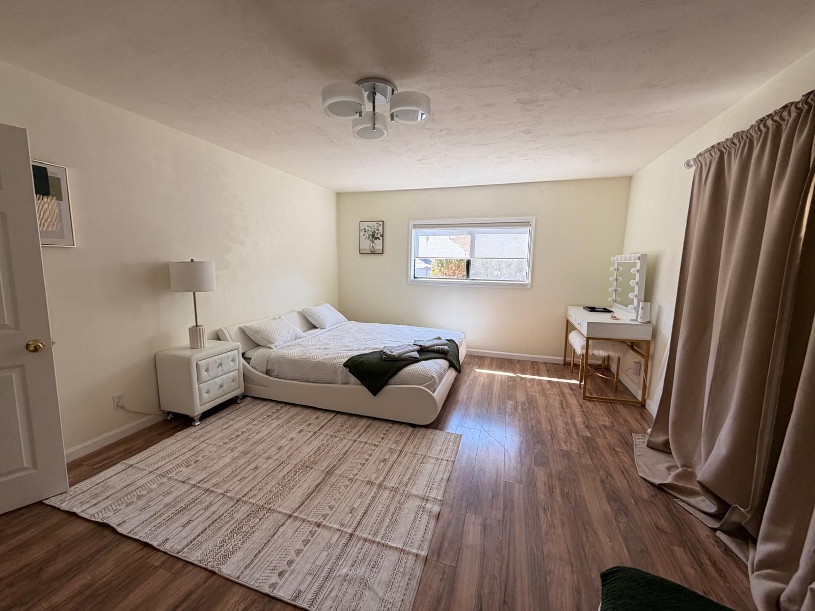Ein Schlafzimmer mit Bett, Nachttisch, Lampe, Fenster und Schminktisch mit Spiegel.