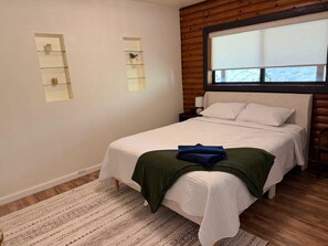 Un dormitorio con cama, mesita de noche, ventana y estante en la pared con una planta.