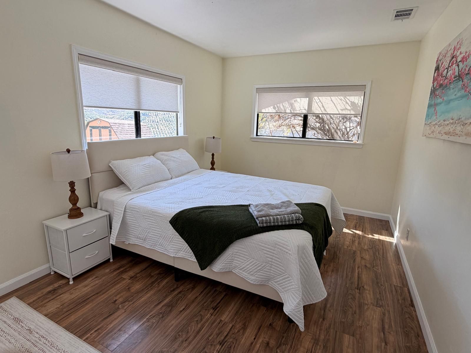 A bedroom with a bed, nightstand, lamp, and two windows.