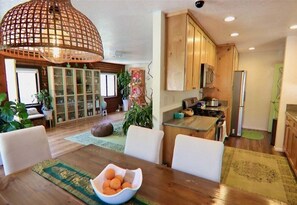 Un comedor con una mesa de madera, sillas y un bol de frutas. Hay una cocina con armarios de madera y un refrigerador.