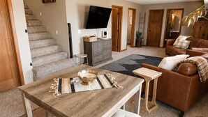 Un vaste espace de vie comprenant une table à manger en bois, des chaises blanches rembourrées, un canapé en cuir marron et un téléviseur à écran plat fixé au mur.