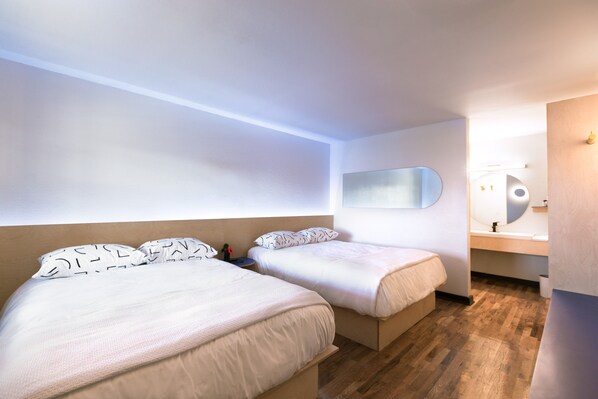 Ett modernt hotellrum med två sängar, en stor spegel, ett trägolv och ett badrum med handfat och spegel.