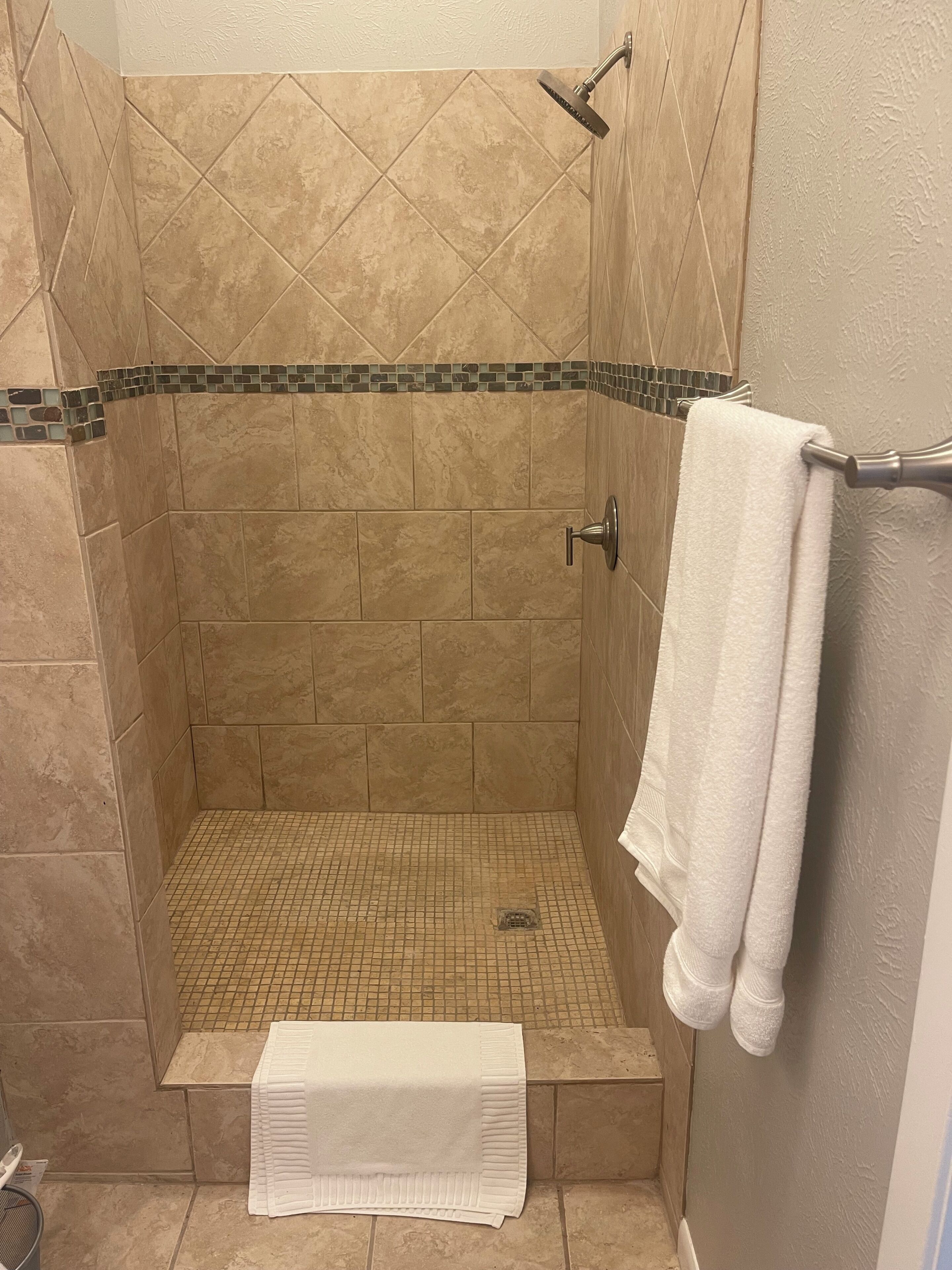 Ducha con azulejos color beige, piso pequeño de azulejos y una toalla blanca en un perchero.