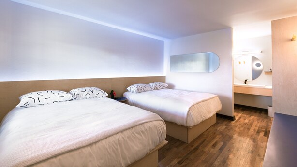 Una habitación de hotel moderna con dos camas, un espejo grande, suelo de madera y un baño con lavamanos y espejo.