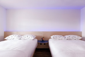 A hotel room with two beds, a small table with a vase, and a plain wall.